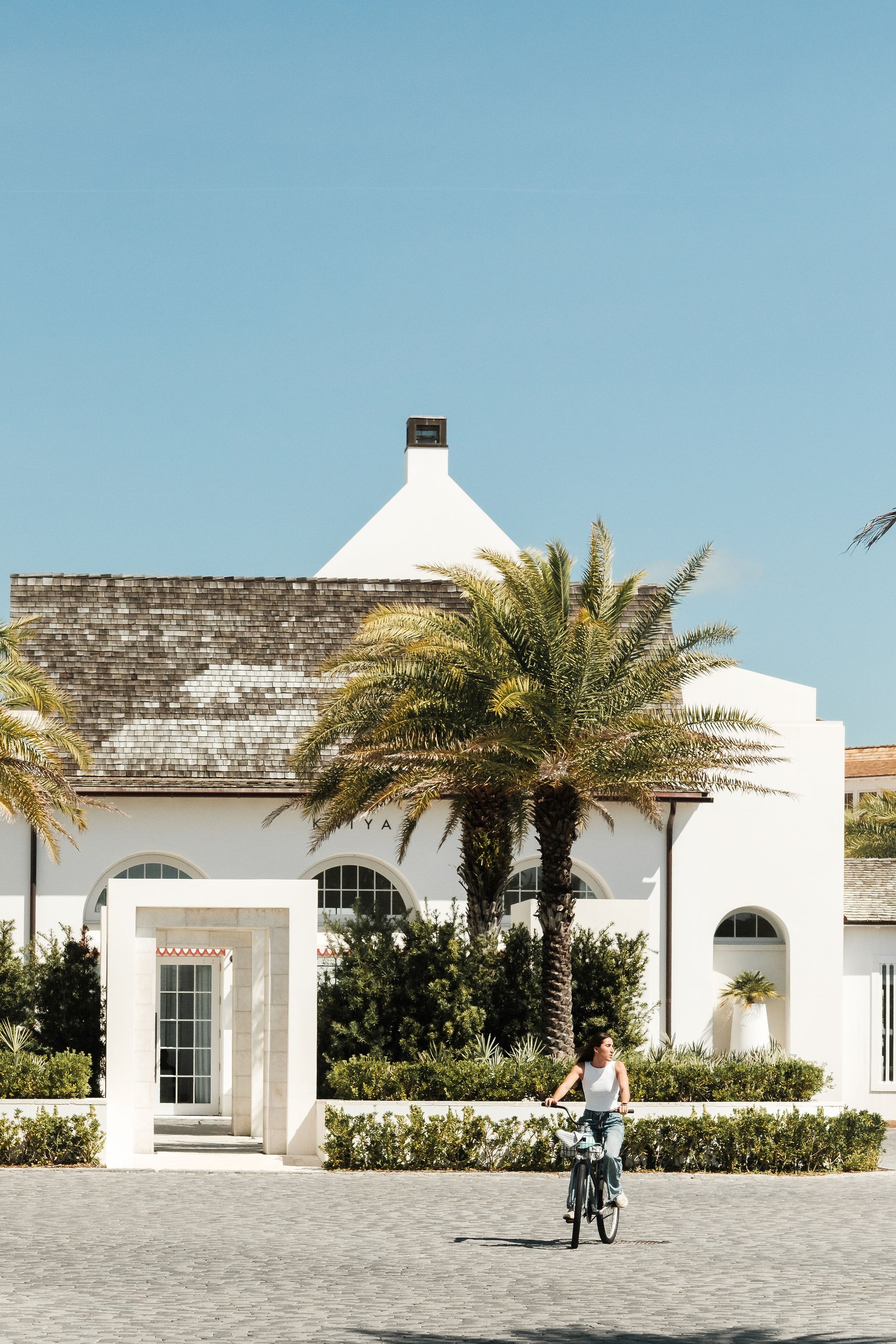 A cyclist rides past a white coastal building framed by palm trees